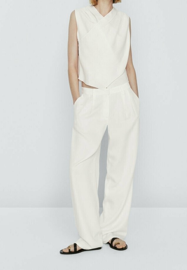 Femme portant un top blanc sans manches drapé et un pantalon large blanc assorti avec des sandales plates noires, mains dans les poches, debout devant un fond uni.