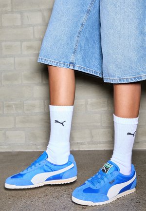 Niebieskie sportowe buty zamszowe i satynowe z białymi akcentami, noszone z białymi prążkowanymi skarpetkami z czarnym logo, zestawione z dżinsowymi szortami.