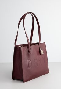 Burgundowa skórzana torba na ramię z wytłoczonym logo "DKNY", z dwoma długimi uchwytami i małym detalem w postaci kłódki przy zamku. Teksturowana powierzchnia.