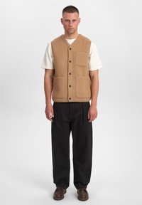 Gilet di lana beige con scollo a V, quattro tasche frontali e bottoni neri, indossato sopra una t-shirt bianca e abbinato a pantaloni neri.