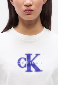 Λευκό βαμβακερό t-shirt με μεγάλο μοβ λογότυπο "CK" και γκρι κείμενο "Calvin Klein" από κά τω. Ριμπ λαιμόκοψη και κοντά μανίκια.