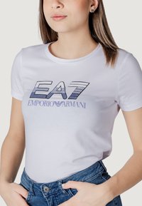 T-shirt bianca con logo "EA7 EMPORIO ARMANI" in blu e nero. Maniche corte, scollatura rotonda, texture in cotone morbido, design aderente.