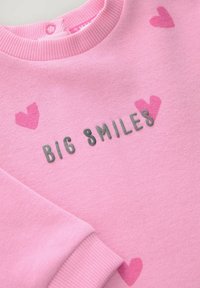 Sweatshirt rose avec un col et des poignets côtelés. Présente le texte "BIG SMILES" en gris et des motifs de cœurs rose dispersés. Texture douce.
