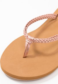 Rosen-guld flätad rem flip-flop med en brun strukturerad sula, som har en thong-stil design och en slät yta.