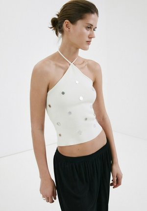 Mujer de cabello oscuro recogido en un moño bajo, vestida con un top halter blanco con adornos circulares plateados y pantalones negros sueltos, sobre un fondo liso.