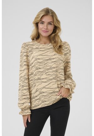 Helles beige Langarmbluse mit wellenförmigem schwarzen Muster, rundem Ausschnitt und gerafften Manschetten. Kombiniert mit schwarzen Hosen. Weiche Stofftextur.