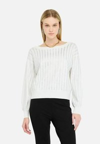 Donna bionda che indossa un maglione bianco a maglia con maniche a palloncino e pantaloni neri, in piedi davanti a uno sfondo bianco.