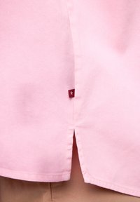 Rosa Baumwollhemd mit abgerundetem Saum und Seitenschlitzen. Verfügt über ein kleines rotes Etikett mit einem weißen Stern nahe der unteren Naht.