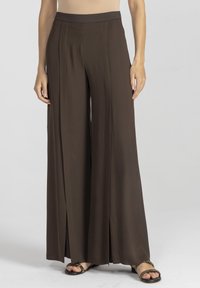 Szerokie spodnie w głębokim brązowym kolorze, wykonane z gładkiego materiału, z wysokim stanem i rozcięciami z przodu dla większej swobody ruchów. W zestawieniu z sandałami na paski.