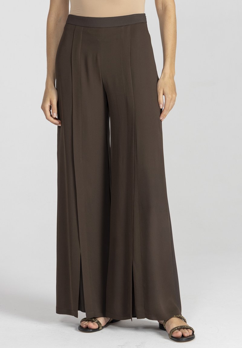 Szerokie spodnie w głębokim brązowym kolorze, wykonane z gładkiego materiału, z wysokim stanem i rozcięciami z przodu dla większej swobody ruchów. W zestawieniu z sandałami na paski.