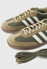 Oliivinvihreät lenkkarit, joissa on beigeä mokkanahkaa, verkkokangasta, valkoiset raidat ja teksturoitu kumipohja. Mukana on sopivat vihreät nauhat.