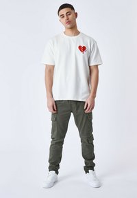 T-shirt bianco a maniche corte con grafica a cuore rosso, abbinato a pantaloni cargo verdi e sneakers bianche, presentato in una posa in piedi.