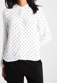 Donna che indossa una blusa bianca a maniche lunghe con pois neri e pantaloni neri, in piedi contro uno sfondo chiaro e uniforme.