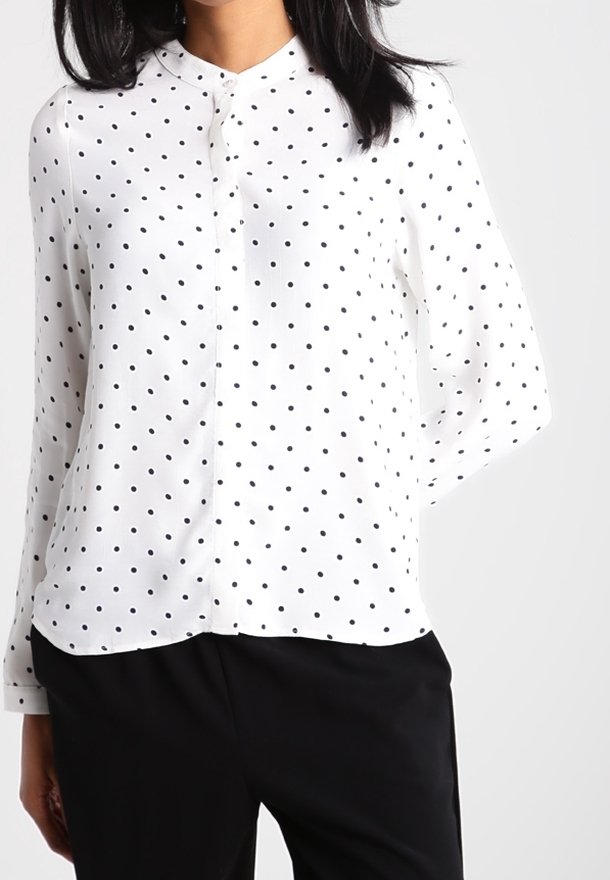 Donna che indossa una blusa bianca a maniche lunghe con pois neri e pantaloni neri, in piedi contro uno sfondo chiaro e uniforme.