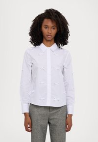White long-sleeve button-up shirt featuring raised white bow accents throughout. Classic collar and smooth cotton material. Textured detail enhances design.