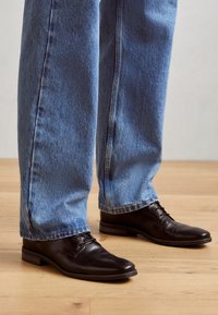 Zapatos de cuero negros con cordones, puntera cuadrada, tacón bajo, llevados con jeans de mezclilla azul holgados, sobre un suelo de madera clara.
