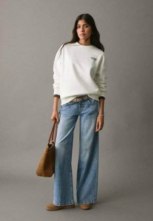 Felpa bianca oversize con logo verde, abbinata a jeans a gamba larga blu chiaro e stivaletti beige in suede, mentre tiene una borsa marrone.
