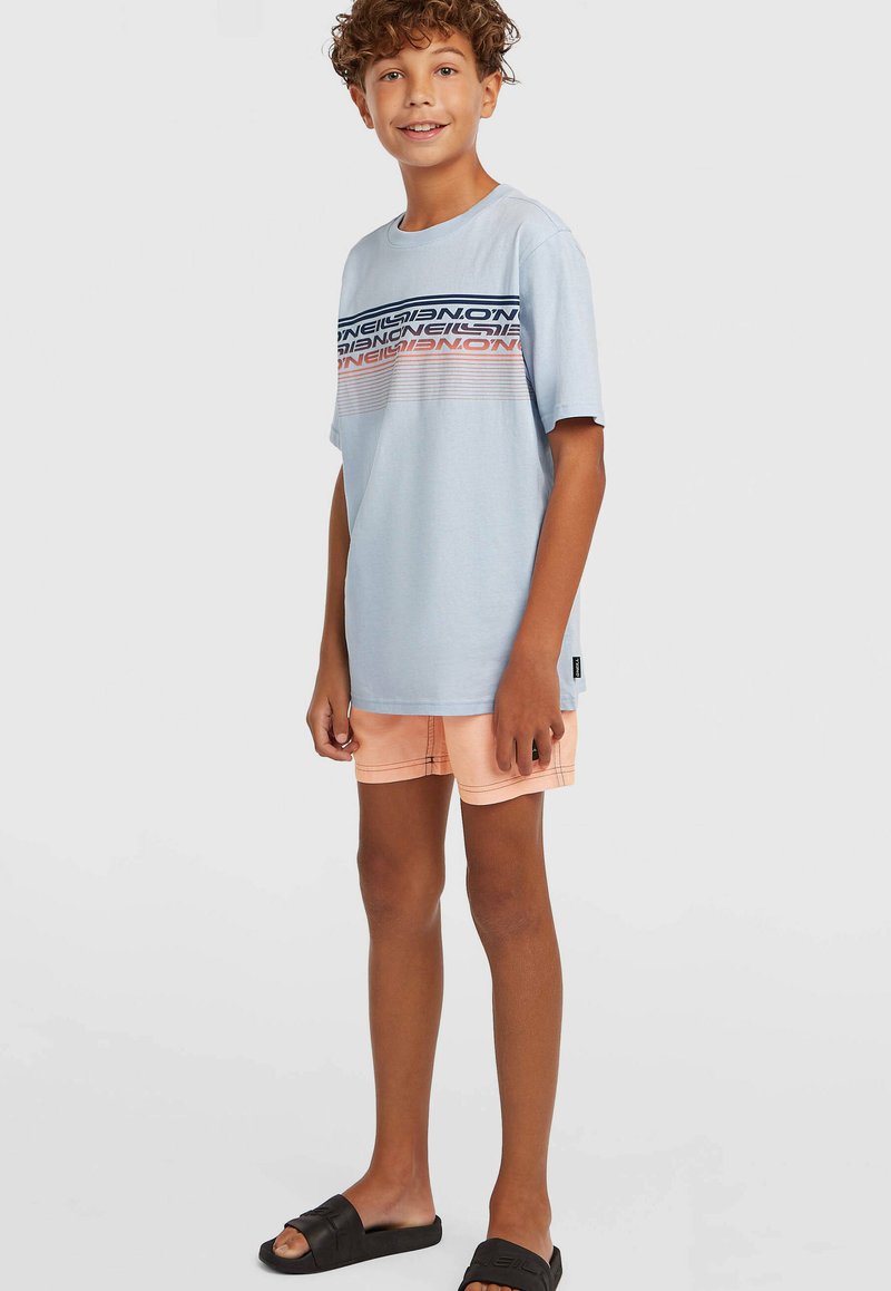 Hellblaues T-Shirt mit einem grafischen Design in Marineblau, Koralle und Weiß; kombiniert mit pfirsichfarbenen Shorts und schwarzen Slidesandalen.