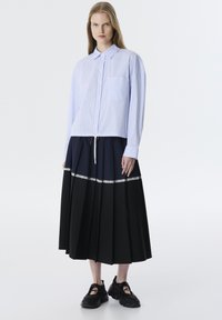 Camisa de botones a rayas azul claro y blanca con mangas largas y bolsillo frontal, combinada con una falda plisada negra con un borde de encaje blanco. Zapatillas negras.