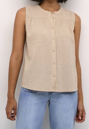 Ærmeløs beige knap-op bluse med lette rynker, båret med lyseblå denimjeans på en person, der står mod en hvid baggrund.