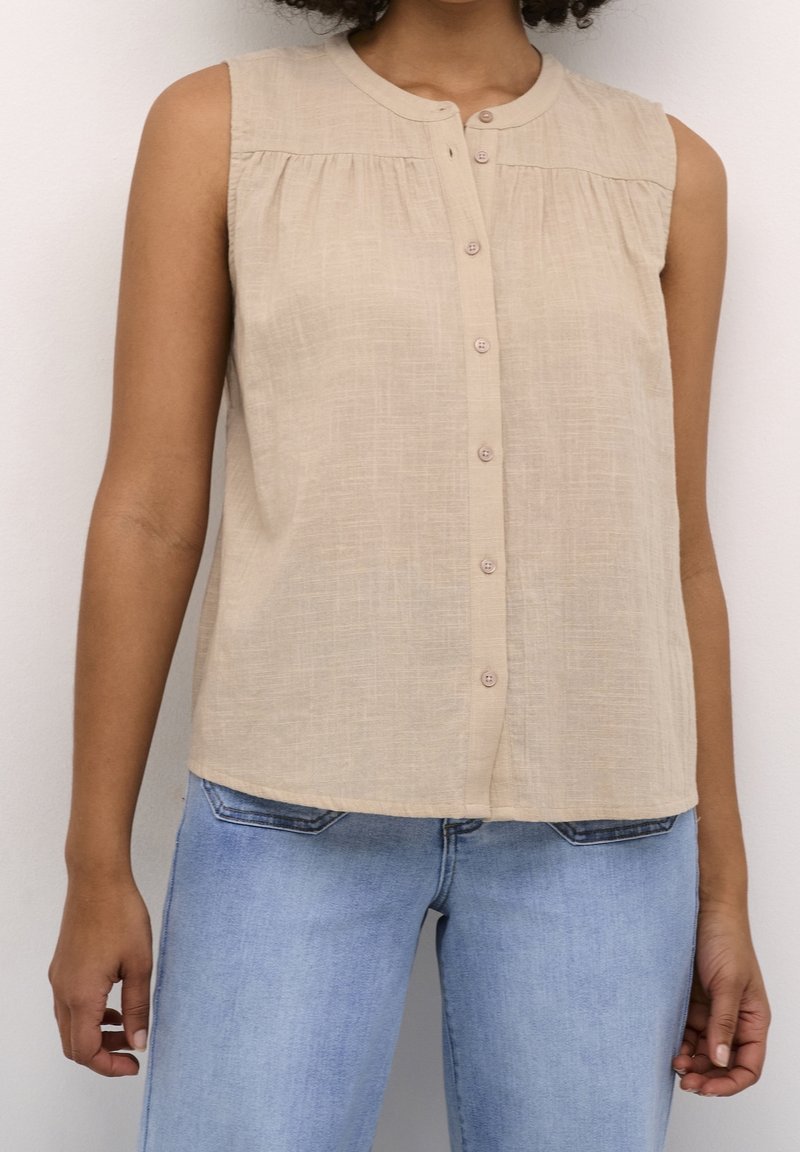 Chemisier beige sans manches boutonné avec de légers fronces, porté avec un jean denim bleu clair sur une personne debout devant un fond blanc.