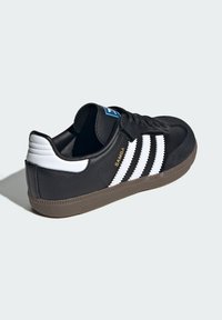 Čierne kožené tenisky s bielymi pruhmi, štruktúrovanou čiernou špičkou a hnedou gumovou podrážkou. Obsahujú modrú nálepku a jemné zlaté branding.