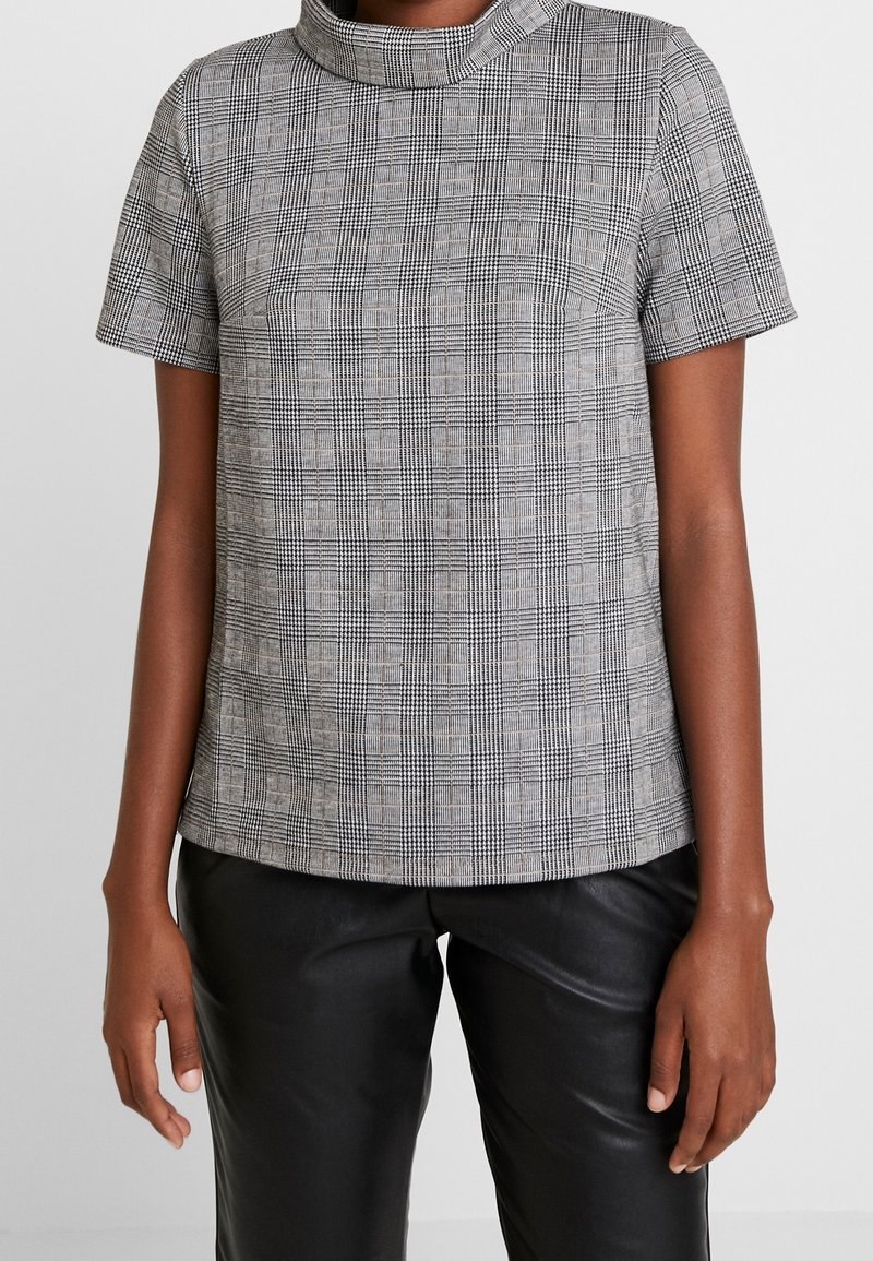 Haut à manches courtes avec un motif à carreaux noir et blanc, présentant un col montant et une coupe structurée, associé à un pantalon en cuir noir.
