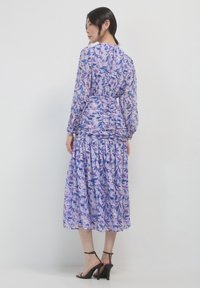 Abito lungo a maniche lunghe di colore viola con stampa floreale, corpino arricciato e gonna a strati, realizzato in un tessuto leggero, abbinato a sandali neri con tacco.