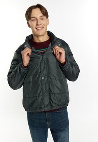 Mørkegrøn pufferjakke med struktureret finish, trykknapper og elastiske manchetter, lagdelt over en stribet sweater, parret med blå jeans.