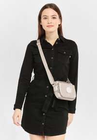Small beige crossbody bag with a smooth texture, featuring a circular accent, adjustable strap, and zipper closure, worn over a black shirt dress.