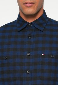 Sinine ja must ruuduline flanellshirt, millel on nööpidega krae, esised taskud ja mustad nööbid; rinnataskul on väike logo.