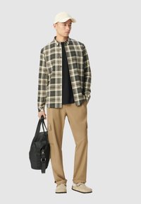 Man in een beige pet, geruite overhemd over een zwarte t-shirt, tan cargo broek, beige schoenen, houdt een zwarte sporttas vast, staande tegen een neutrale achtergrond.
