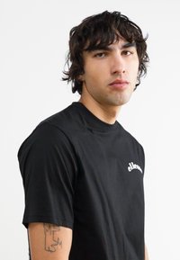 Camiseta negra de cuello redondo que presenta un pequeño logo blanco en el pecho. La tela es suave y tiene mangas cortas, utilizada por un modelo con cabello rizado y oscuro.
