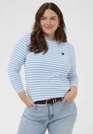 Femeie cu păr lung și castaniu, purtând un pulover cu mâneci lungi în dungi albastre și albe, cu un mic detaliu în formă de inimă, blugi deschis la culoare și o curea închisă la culoare.