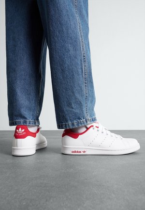 Personne portant des baskets blanches Adidas Stan Smith avec des accents rouges et un jean bleu, debout sur un sol gris devant un fond uni.