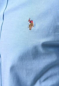 Broderad logotyp av en polo-spelare i röd och blå, ridande på en brun häst, på ljusblått texturerat tyg.