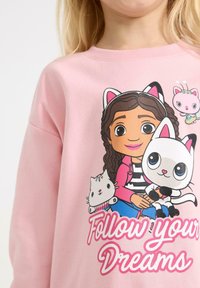 Roze sweatshirt met een illustratie van een meisje met kattenoren, dat een witte kat en een cupcake vasthoudt, met de tekst "Volg je Dromen" eronder.