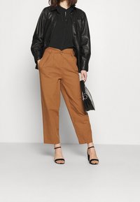 Veste en cuir noire sur un haut sombre, associée à un pantalon taille haute marron et des talons noirs à lanières. Tient un sac à main noir.