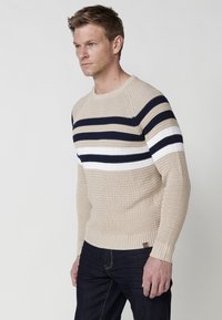 Beige kötött pulóver navy, fehér és krémszínű vízszintes csíkokkal. Kerek nyakkivágással és bordázott mandzsettákkal, valamint alsó résszel rendelkezik.
