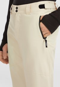 Beige geïsoleerde broeken met een ritszak, drukknoopsluitingen en een klittenbandsluiting. De stof is glad met een iets glanzende afwerking.