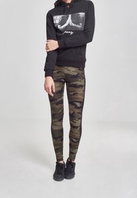 Felpa nera con stampa grafica e testo sul davanti, abbinata a leggings mimetici con motivi verde scuro e nero. Sneakers nere.