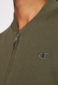 Olivgrüner Reißverschluss-Sweatshirt aus strukturiertem Stoff, mit einem gerippten Kragen und gesticktem Logo auf der linken Brust.