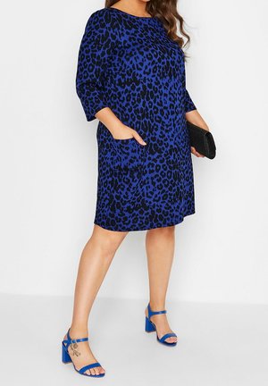 Kobieta ubrana w niebieską i czarną sukienkę w panterkę do kolan z rękawami do połowy ramion, niebieskie sandały na słupku z otwartym noskiem, trzymająca czarną kopertówkę.