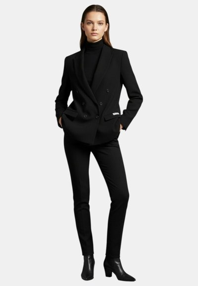 Donna in piedi con le mani in tasca, indossa un blazer doppiopetto nero, un dolcevita nero, pantaloni neri aderenti e stivaletti neri.
