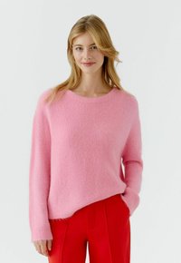 Frau mit blondem Haar, die einen weichen rosa Pullover und leuchtend rote Hose trägt, steht mit einer Hand in der Tasche vor einem weißen Hintergrund.