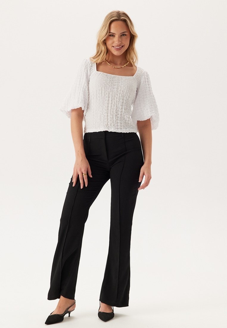 Top bianco texturizzato con scollatura quadrata e maniche a sbuffo corte, abbinato a pantaloni neri a vita alta e tacchi a punta neri.