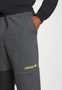 Grå joggers med svart midjeband, som har sidofickor och en gul "adidas"-logotyp på underbenet. Mjuk tyg, avslappnad design.
