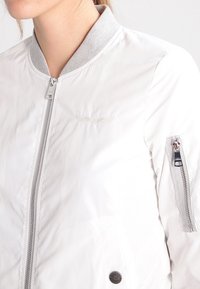 Biała kurtka bomberka z szarym kołnierzem, z zamkiem błyskawicznym z przodu, kieszenią z zamkiem na lewym rękawie oraz haftowanym logo nad klatką piersiową.
