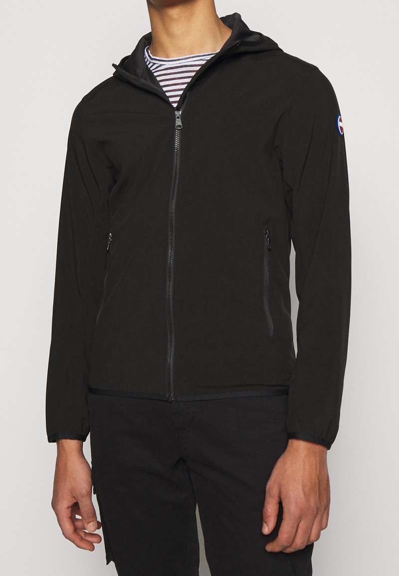 Person in einer schwarzen Reißverschluss-Hoodie-Jacke über einem gestreiften Hemd, mit entspannten Händen an den Seiten, vor einem schlichten Hintergrund.