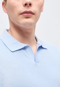 Κοντινό πλάνο στο κατώτερο μέρος του προσώπου και τον λαιμό ενός ατόμου που φοράει ένα ανοιχτό γαλάζιο πικέ πόλο μπλουζάκι με ανοιχτό γιακά μπροστά σε ένα απλό φόντο.